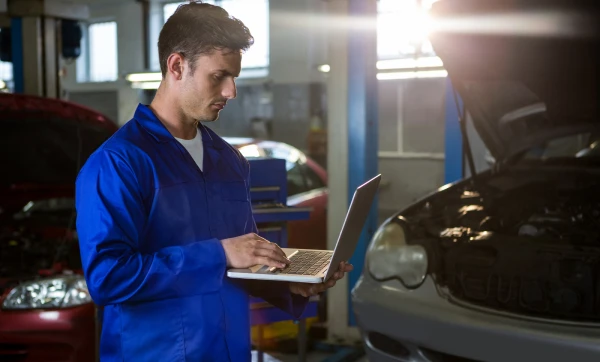 Profissional de mecânica de pé ao lado de um carro em manutenção na oficina usando um notebook para saber Como formar o preço de venda de serviços (mão de obra + custos).