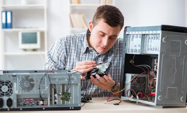 Profissional de TI masculino, sentado fazendo manutenção de reparo em um computador em sua bancada de trabalho representando: Responsabilidade civil do técnico: quando a assistência técnica responde por dano ao equipamento.