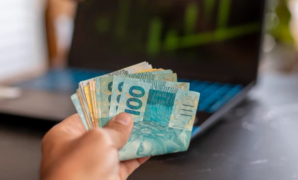 uma mão com várias notas de 100 reais em frente a um notebook, representando: 13º salário: como calcular, quando pagar e o que muda para quem foi admitido no meio do ano