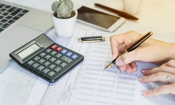 Uma mão com uma caneta escrevendo em um documento ao lado de uma calculadora, representando: Folha de pagamento para pequenas empresas: o que compõe, como calcular e o papel do ERP