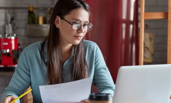 Mulher de óculos segurando um documento enquanto utiliza um notebook, representando a análise de curva ABC de produtos no Gestão Click 1x.