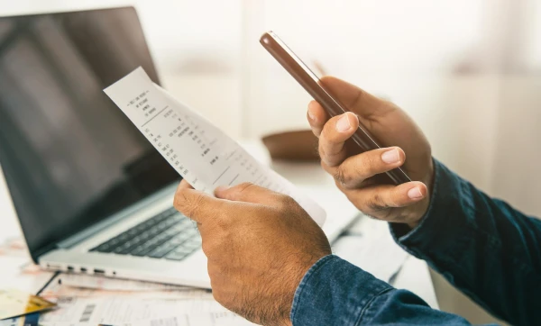 Mãos segurando um celular e um papel em frente a um notebook, representando: Como emitir boleto bancário pelo GestãoClick: passo a passo completo