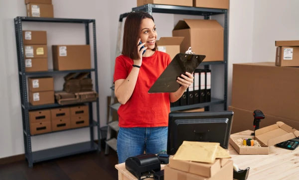 Mulher com prancheta em mão falando ao telefone, representando: Como configurar estoque mínimo e alertas de reposição no GestãoClick title: Como Configurar Estoque Mínimo e Alertas de Reposição